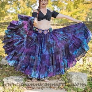 Op de afbeelding: Een vrouw in een zwarte crop top en een gelaagde rok met een tie-dye patroon in paars en blauw. De rok wordt vastgehouden om zijn volle vorm te laten zien. Een decoratieve riem met bloemaccenten zit om de taille. De achtergrond is een park.