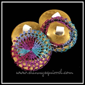 May include: Two gold cymbals with crocheted covers. The covers are a mix of purple, blue, and teal colors. The cymbals have a decorative design and are held together with a black elastic band.