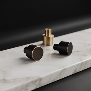 May include: Three cylindrical cabinet knobs, two with a matte black finish and a thin gold-toned rim, and one central knob featuring a solid brushed gold-toned finish. Each knob has a flat circular face and a smaller cylindrical base for mounting.