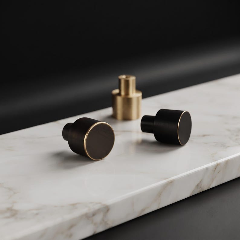 May include: Three cylindrical cabinet knobs, two with a matte black finish and a thin gold-toned rim, and one central knob featuring a solid brushed gold-toned finish. Each knob has a flat circular face and a smaller cylindrical base for mounting.