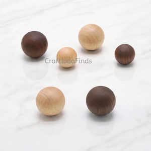 Pode incluir: Cinco puxadores esféricos de madeira em vários tamanhos e tons de madeira, variando de madeira natural clara a madeira castanha escura. O texto CraftivaaFinds está impresso no centro do arranjo. Estes objetos redondos e lisos estão agrupados.