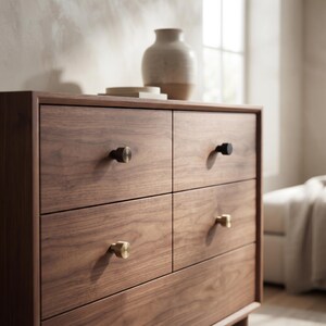 May include: A wooden dresser featuring four drawers with a visible grain pattern. Each drawer is equipped with a single knob, including two metallic gold-toned knobs, one matte black knob, and one brushed brass knob, all with a cylindrical shape.