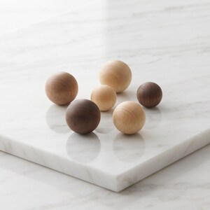 May include: A collection of six smooth, spherical wooden balls in varying sizes and natural wood tones, ranging from light beige to dark brown. The spheres are arranged in a cluster, showcasing distinct wood grain patterns on their surfaces.