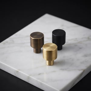 May include: Three cylindrical cabinet knobs with a smooth, solid finish. One knob is finished in a brushed gold tone, one in a matte black, and one in a dark bronze. Each knob features a flat circular top and a smaller cylindrical base.