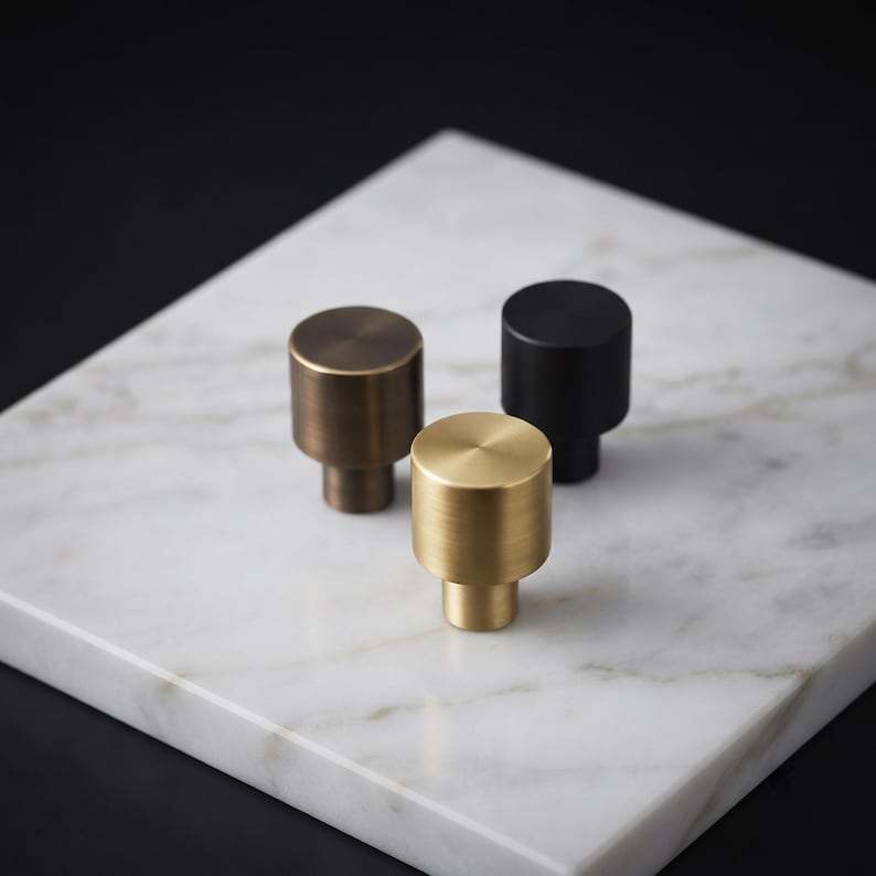 May include: Three cylindrical cabinet knobs with a smooth, solid finish. One knob is finished in a brushed gold tone, one in a matte black, and one in a dark bronze. Each knob features a flat circular top and a smaller cylindrical base.
