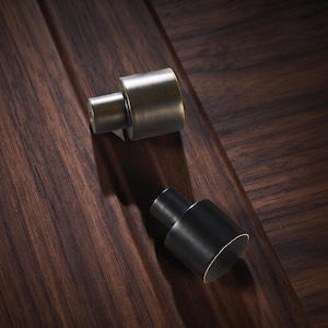May include: Two cylindrical cabinet knobs, one with a brushed antique brass finish and the other with a matte black finish. Each knob features a stepped design with a smaller circular base and a wider cylindrical top, showcasing a clean profile.