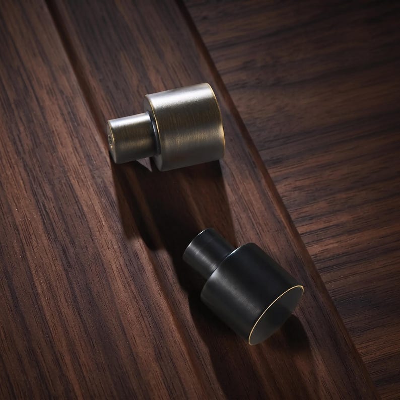 May include: Two cylindrical cabinet knobs, one with a brushed antique brass finish and the other with a matte black finish. Each knob features a stepped design with a smaller circular base and a wider cylindrical top, showcasing a clean profile.