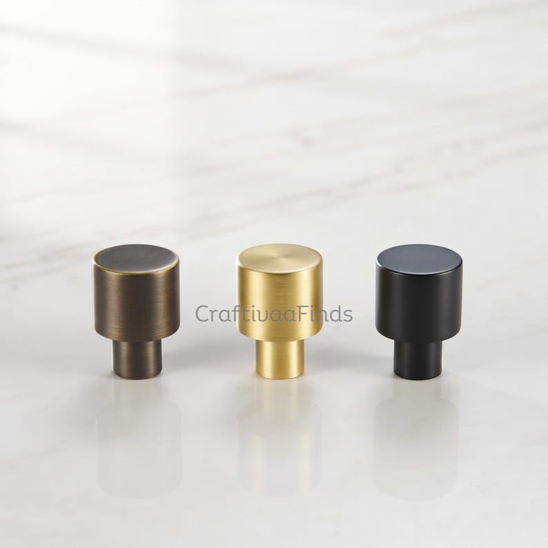 May include: Three cylindrical cabinet knobs arranged in a row. The left knob is dark bronze, the center knob is brushed gold, and the right knob is matte black. Each knob features a flat circular top and a smaller cylindrical base for mounting.