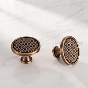 May include: Two round cabinet knobs with a brushed bronze finish. Each knob features a flat circular face with a recessed center containing a dark, intricate, repeating geometric lattice pattern. The knobs have a smooth, curved outer edge.