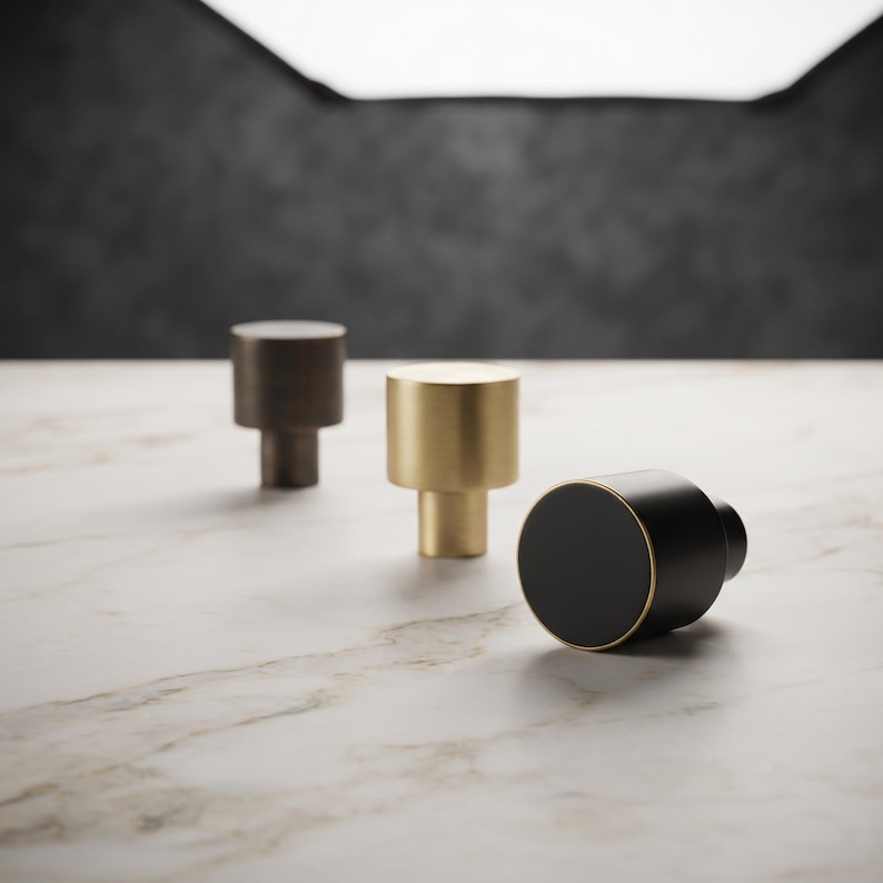 May include: Three cylindrical cabinet knobs arranged in a row. One knob is dark bronze, one is brushed gold, and the third is matte black with a thin gold-toned rim. Each knob features a flat circular top and a smaller cylindrical base.