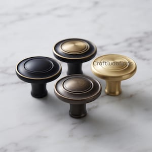 May include: Four round cabinet knobs with a stepped circular design. One is matte black, one is brushed gold, one is a two-tone black and gold, and one is a dark bronze finish. The text Craftivaafinds is visible on the gold knob surface.