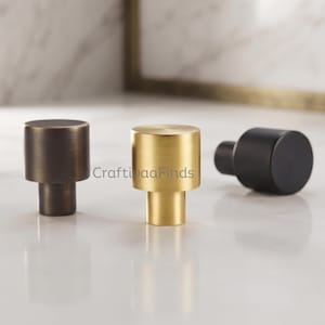 May include: Three cylindrical cabinet knobs in different finishes: one dark bronze, one brushed gold, and one matte black. Each knob features a flat circular top and a smaller cylindrical base. The text CraftivaaFinds is visible in the center.