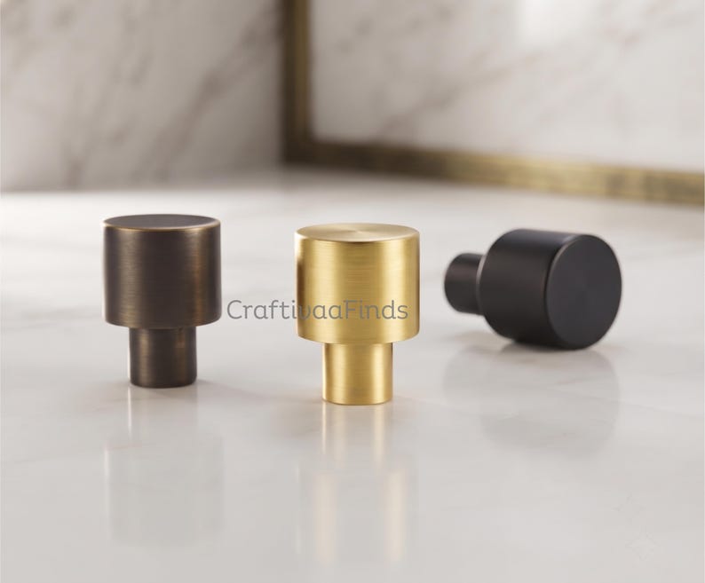 May include: Three cylindrical cabinet knobs in different finishes: one dark bronze, one brushed gold, and one matte black. Each knob features a flat circular top and a smaller cylindrical base. The text CraftivaaFinds is visible in the center.
