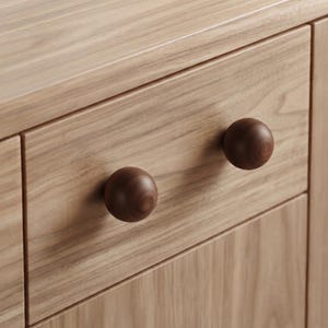 May include: A light-toned wooden drawer front featuring two dark brown, spherical wooden knobs. The drawer is constructed from wood with a visible grain pattern, and the two round knobs are mounted horizontally on the flat surface of the drawer.