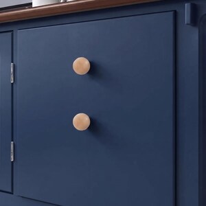 May include: Two round, light-colored wooden knobs are mounted vertically on a flat, dark navy blue cabinet door. The knobs have a smooth, spherical shape and a natural wood grain finish, contrasting against the solid matte blue surface.