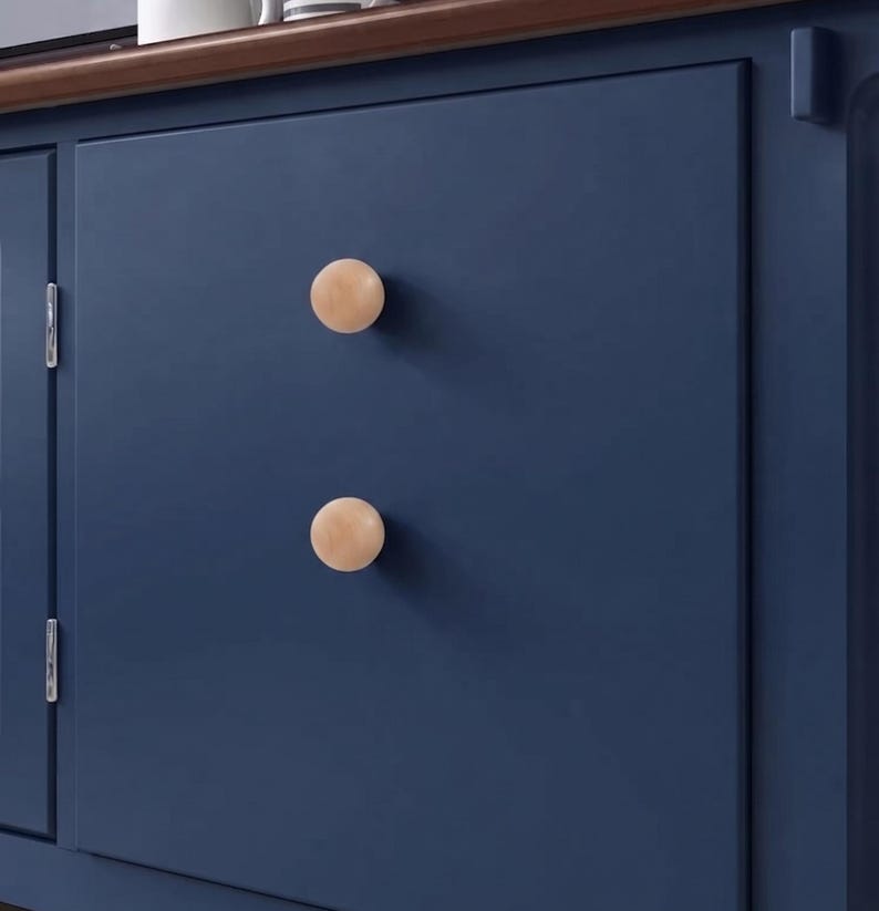 May include: Two round, light-colored wooden knobs are mounted vertically on a flat, dark navy blue cabinet door. The knobs have a smooth, spherical shape and a natural wood grain finish, contrasting against the solid matte blue surface.