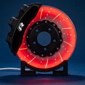 Op de afbeelding: Een ronde remschijflamp met een gloeiend rood lichteffect, voorzien van een zwarte remklauw met een wit R-logo. Het geheel is gemonteerd op een zwarte standaard en bevat een wit netsnoer dat uit de basis van de eenheid komt.
