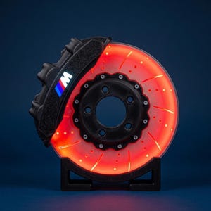 May include: A glowing red LED brake disc lamp featuring a black brake caliper with the M logo in blue, purple, and white. The circular disc has drilled holes and curved ventilation slots, mounted on a black stand against a dark blue background.