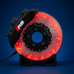 Peut inclure: Une lampe en forme de disque de frein automobile avec un étrier noir portant l'inscription RS en blanc. La surface du disque est éclairée par une lueur rouge vive, mettant en évidence les trous percés et les fentes de ventilation incurvées.