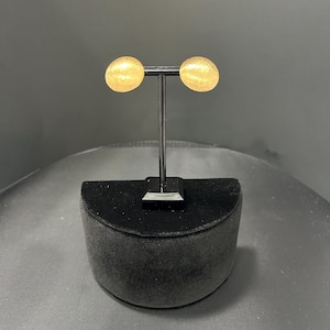 May include: A pair of gold-colored stud earrings with a textured surface. The earrings are round and displayed on a black metal stand with a black velvet base. The background is a dark gray.