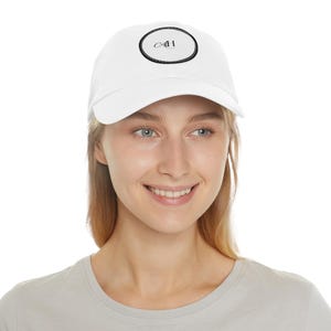 Könnte beinhalten: Weiße Baseballkappe mit gebogenem Schirm. Die Kappe hat ein schwarzes ovales Design mit den Buchstaben "CAI" in der Mitte. Die Kappe wird von einer Person getragen.