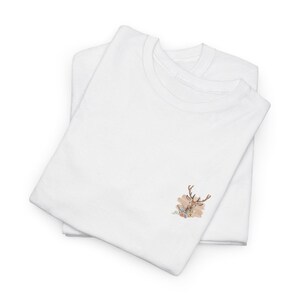 May include: Two white t-shirts are stacked, with a deer head and floral design embroidered on the front. The deer is brown with antlers, and the flowers are in shades of pink, peach, and blue. The shirts are folded neatly.