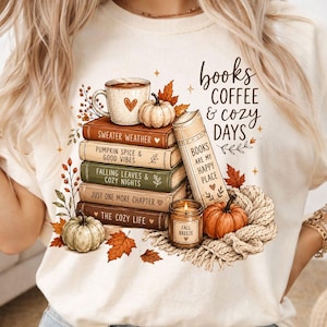 May include: A cream-coloured t-shirt featuring a graphic of a stack of five books, a coffee mug, two pumpkins, a lit candle labelled Fall Breeze, and a knit blanket. The text reads Books Coffee & Cozy Days and Books Are My Happy Place.