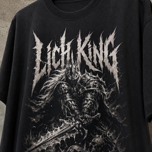 Puede incluir: Una camiseta negra de manga corta con un estampado gráfico de una figura oscura con armadura, una corona de pinchos y una espada dentada. El texto LICH KING aparece impreso en una fuente blanca desgastada sobre la figura central.