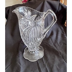 May include: Clear glass pitcher with intricate starburst and geometric patterns. The pitcher features a scalloped rim, a sturdy handle, and a decorative base. A classic piece for serving beverages.