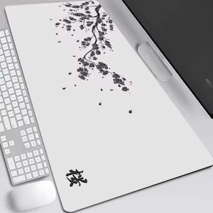 Op de afbeelding: Witte rechthoekige bureaumat met een zwart bloemen takontwerp. Het ontwerp omvat een tak met bladeren en vallende bloemblaadjes. Een toetsenbord en muis zijn zichtbaar aan de linkerkant. De mat heeft ook een Japans karakter.