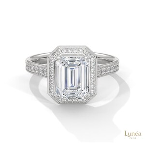 May include: A silver engagement ring with a large emerald-cut diamond center stone. The center stone is surrounded by a halo of smaller diamonds. Additional diamonds are set along the band. The ring is set against a white background.