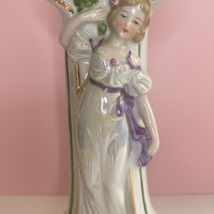 Może przedstawiać: Porcelanowa figurka kobiety w powłóczystej białej sukni, ozdobionej fioletową szarfą. Figurka trzyma się kolumny z kompozycją kwiatową na górze, z fioletowymi kwiatami i zielonymi liśćmi. Złote akcenty zdobią krawędzie.