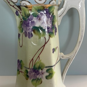 May include: Antique porcelain coffee pot with a lid, featuring hand-painted purple violets and green leaves on a pale yellow background. The handle and spout are white, and the rim has a blue and gold trim. A decorative piece.