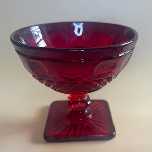 May include: A ruby red glass dessert dish with a square base and a round bowl. The bowl has a decorative pattern around the rim and a textured design on the lower portion. The stem is short and the base is square.