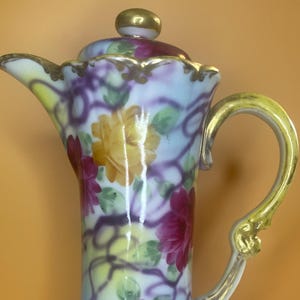 Könnte beinhalten: Eine Vintage-Porzellankanne mit floralem Design. Die Kanne hat einen hellblauen Hintergrund mit gelben, rosa und lila Blumen und Ranken. Der Griff und der Rand sind mit goldenen Zierleisten versehen. Die Kanne hat einen Deckel mit einem goldenen Knopf.