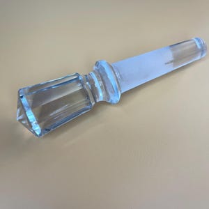 May include: A clear glass bottle stopper with a faceted, geometric handle and a frosted, tapered body. The stopper is designed to fit into a bottle opening. The glass has a slight blue tint.