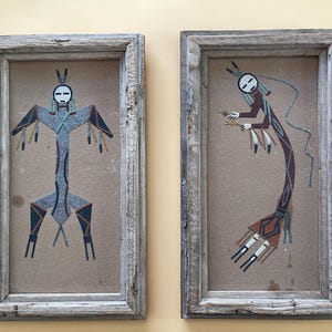 May include: Two rectangular wall art pieces featuring sand paintings of stylised figures in weathered wooden frames. The left figure is symmetrical with blue and grey tones, while the right figure is curved with brown, red, and white accents.