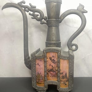 May include: Antique metal pipe with ornate dragon and bird details. The central body features three panels with painted scenes of figures and landscapes. The pipe has a curved stem and a decorative top.