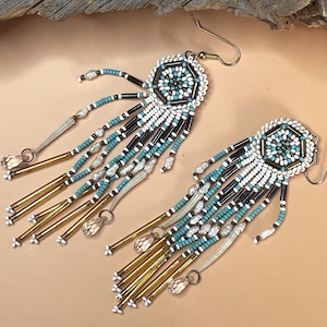 May include: A pair of long beaded earrings featuring a hexagonal top section in white, turquoise, and black. The design includes dangling fringe strands made of white, turquoise, and black seed beads, long metallic gold tubes, and clear crystal drops.