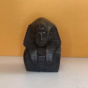 May include: A dark, carved bust of an Egyptian pharaoh, featuring a detailed headdress and serene facial expression. The sculpture is a dark, possibly black, color and sits on a white surface against an orange background. The bust is a decorative item.