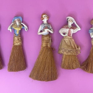 May include: A collection of seven decorative figurines featuring porcelain torsos of figures wearing various outfits, each attached to a long, brown, fibrous broom-like skirt. The figures are arranged in a row, displaying diverse clothing styles.