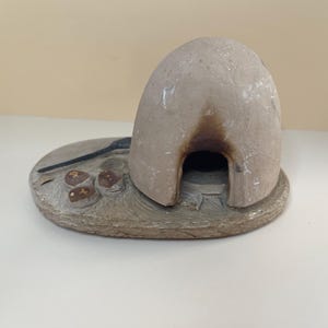 May include: A beige, dome-shaped clay oven sculpture set on an oval base. The structure features a dark, scorched arched opening at the front. Beside the oven, three small brown circular objects and a thin black tool are molded onto the base.