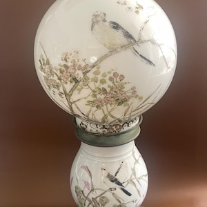 May include: A white glass lamp with a spherical shade and a smaller base, both decorated with painted birds and floral branches. The lamp features a metallic, weathered green-toned base with ornate detailing and a matching metal collar section.