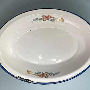 May include: An oval enamel basin with a white interior and exterior, featuring a dark blue rim. The basin is decorated with floral motifs in shades of pink, yellow, and green at the top and bottom. There is visible wear and chipping on the rim.