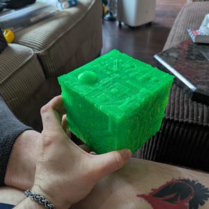 May include: A bright green, translucent, three-dimensional cube featuring intricate, raised geometric patterns and mechanical-looking surface details across its faces. The object has a textured, industrial appearance with a circular indentation on one side.