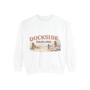May include: A white long-sleeve sweatshirt featuring a graphic print on the chest. The print displays the text DOCKSIDE DARLING in red and black lettering above a watercolor-style illustration of a beach scene with wooden posts and flowers.