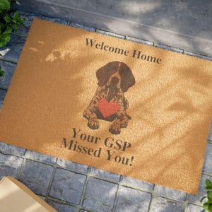May include: A rectangular tan coir doormat featuring a brown German Shorthaired Pointer illustration with a red heart on its chest. The text Welcome Home is printed above the dog, and the text Your GSP Missed You! is printed below the dog.