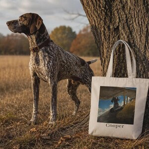 Pode incluir: Uma sacola de lona branca com uma estampa gráfica retangular de um cão sentado na cabine de um pequeno avião. Abaixo da imagem, o nome Cooper está impresso em texto preto. A bolsa tem duas alças superiores e um formato retangular.