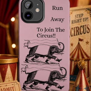 以下が含まれることがあります： サーカスをテーマにしたピンク色の携帯電話ケース。ケースには「Run Away To Join The Circus!!」というテキストと象のイラストが描かれています。ヴィンテージのサーカスのチケットとパーティーハットも表示されています。