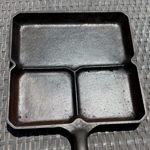 May include: A black cast iron skillet featuring a rectangular design with three distinct cooking compartments. The pan includes one large rectangular section at the top and two smaller square sections at the bottom, complete with a sturdy handle.
